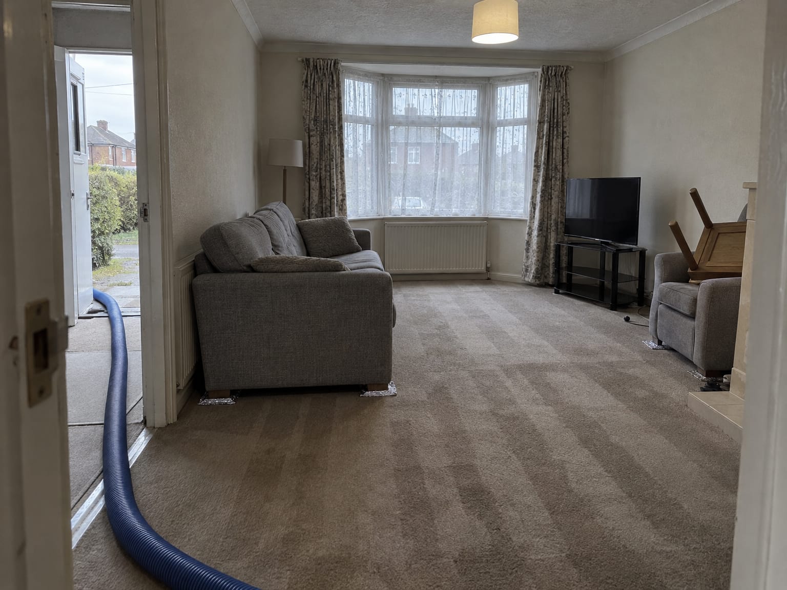 An empty living room with a freshly cleaned damp carpet, the sofa on foil squares and a hose through the doorway