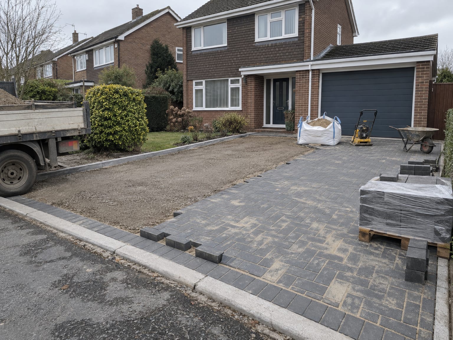 A half-laid block paved drive with a pallet of blocks, a bag of sand and a plate compactor by the garage